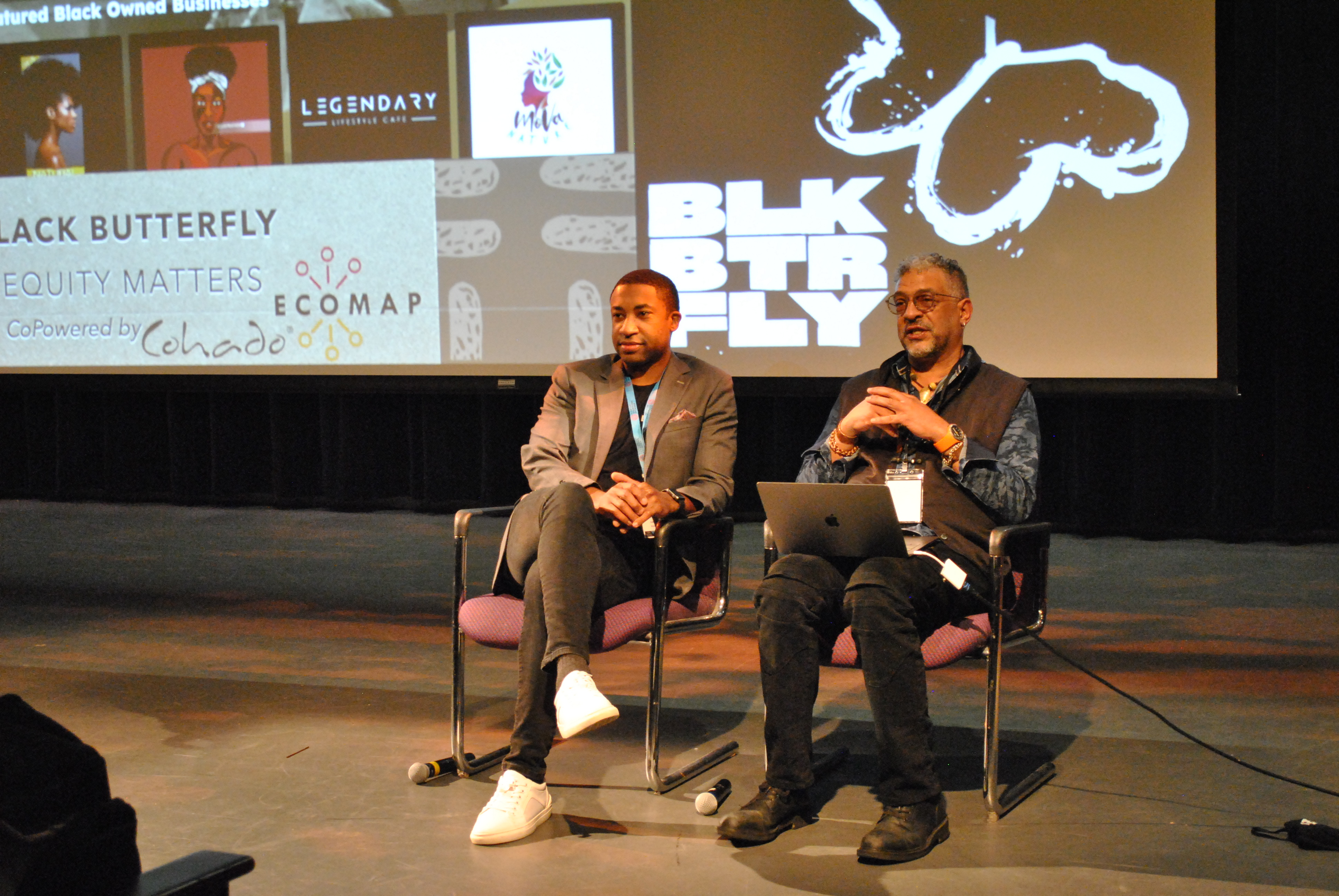 A photo of two black men, Sherrod Davis and Paulo Gregory, sitting on a stage with a projection screen behind them