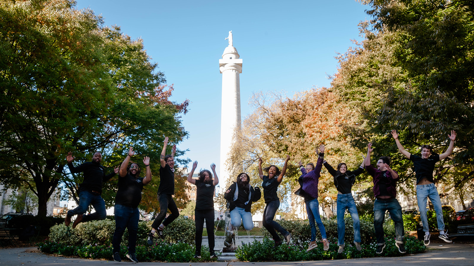 A group of young people in black shirts jumping in front a park with a large white monument